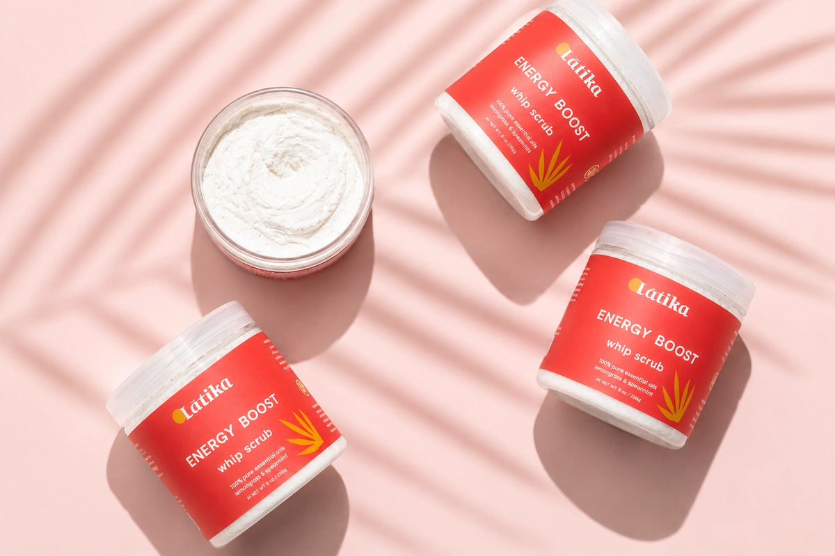 Four jars of Latka Energy Boost Whip Scrub arranged on a pink background with soft leaf shadows. One jar is open, showing the white whipped scrub inside, while the others are closed with bright red labels and white caps.