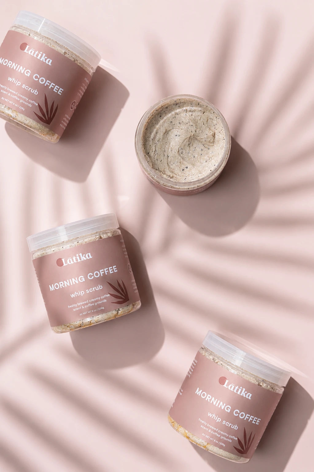 Three Latika Morning Coffee whipped scrub jars and one open jar showing the cream product, arranged on a pink background with leaf shadow overlays.