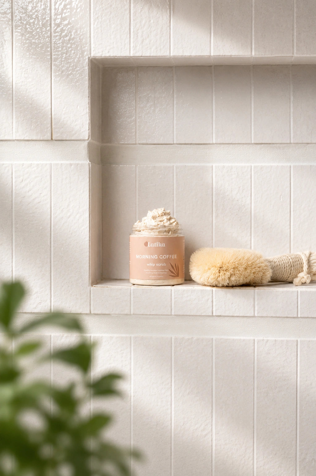 A jar of Bath & Body Works Whiskering Cream sits on a white tiled shelf next to a natural bristle brush and a cotton washcloth, with soft sunlight and a blurred green plant in the foreground.