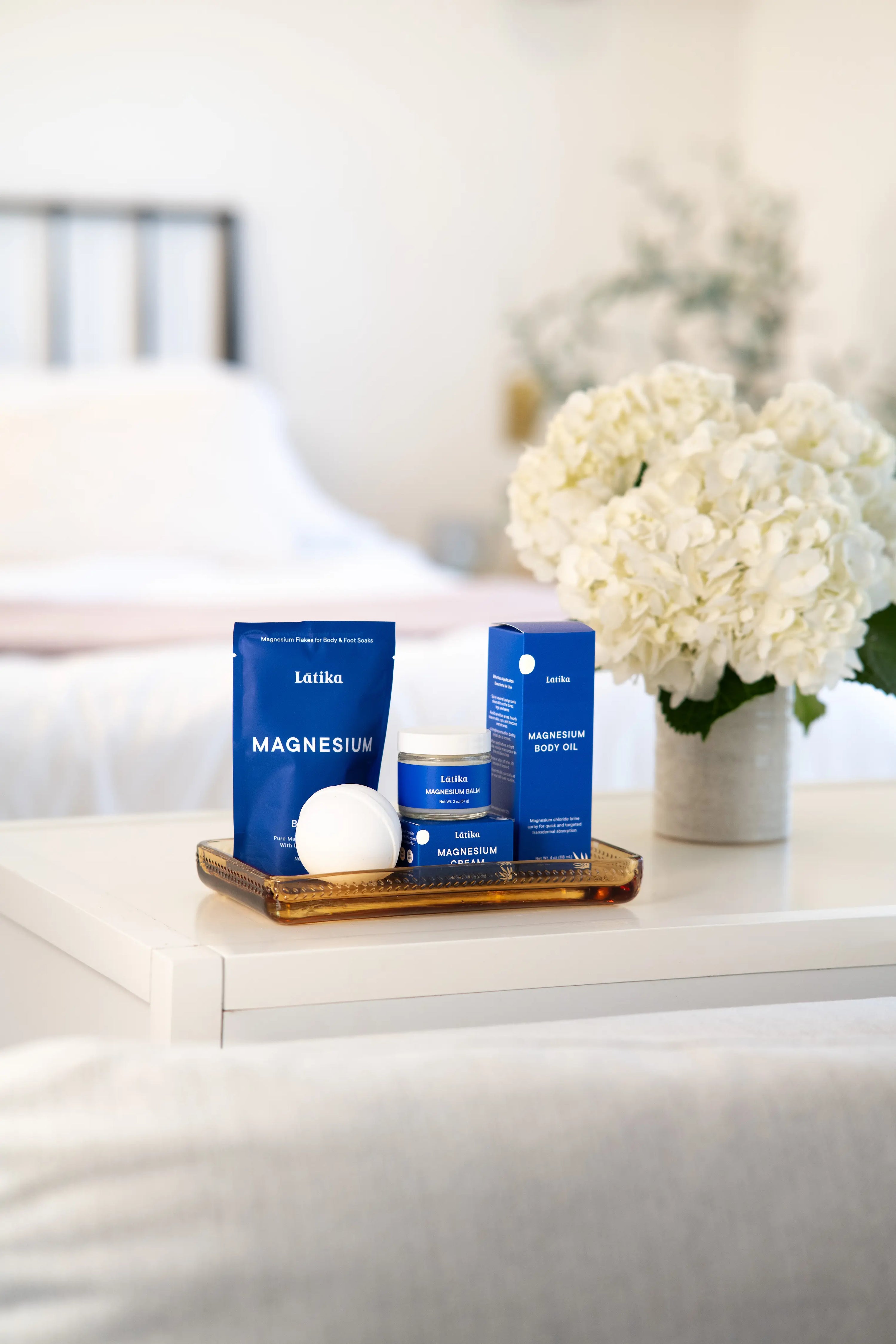 Magnesium products on a tray in a bedroom. including magnesium soak, magnesium cream and balm, magnesium oil, and a bath bomb. All in blue packaging by Latika Beauty
