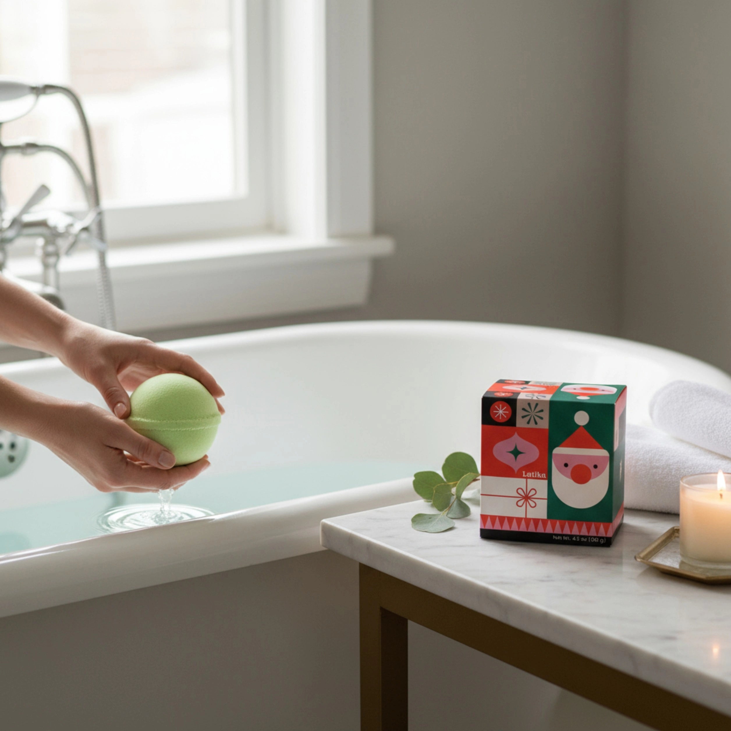 Person holding a green bath bomb next to a bathtub with a decorative box and candle on a nearby surface.