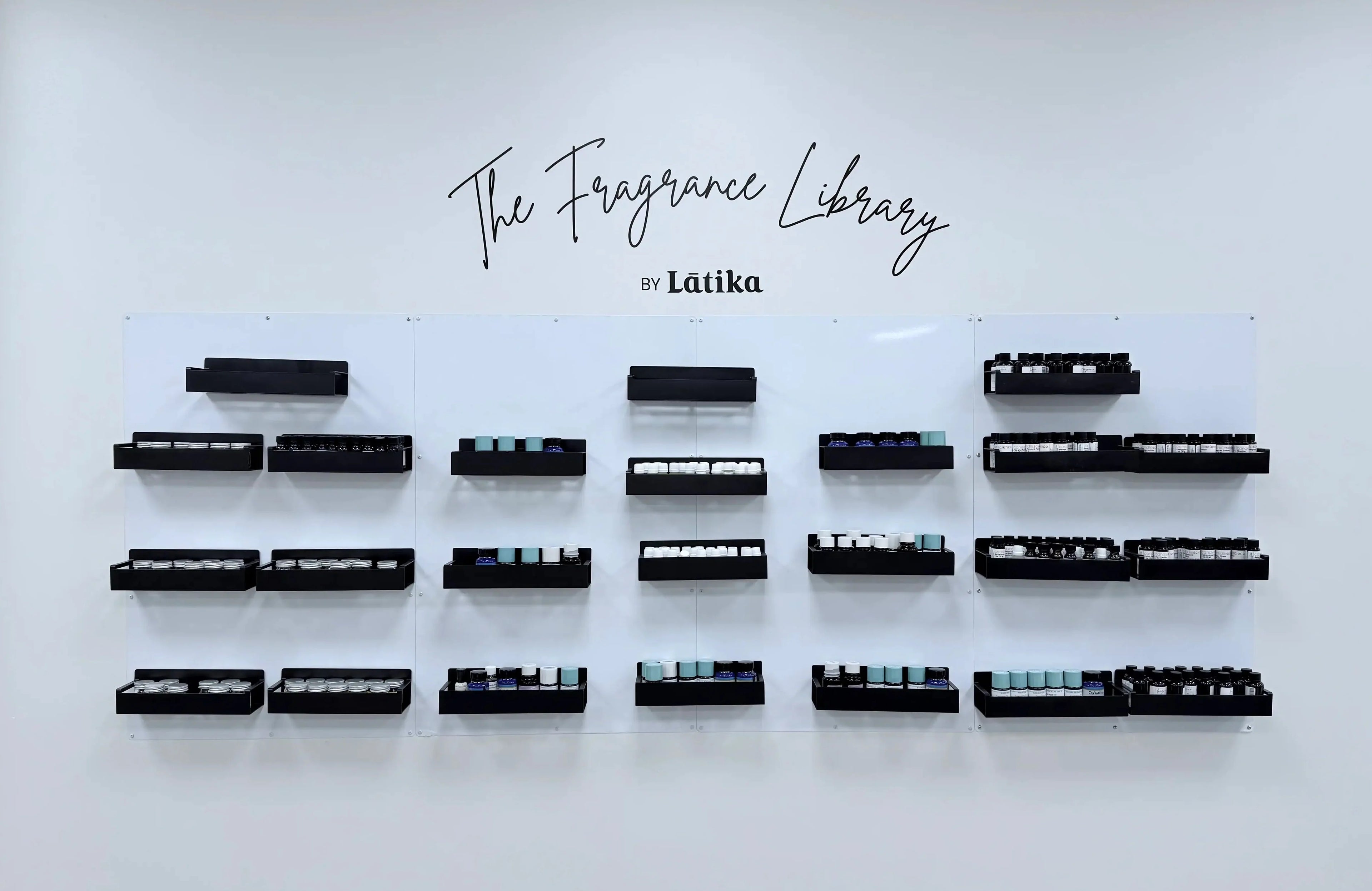 Wall-mounted display of black fragrance bottles labeled 'The Fragrance Library by Látika' on a white background.