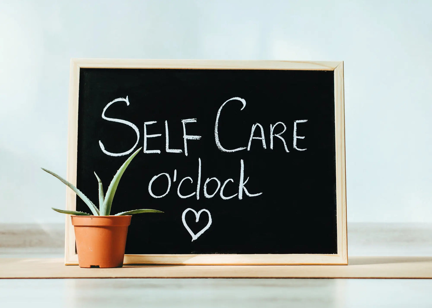 Blackboard with 'Self Care o'clock' text and a potted plant on a light background