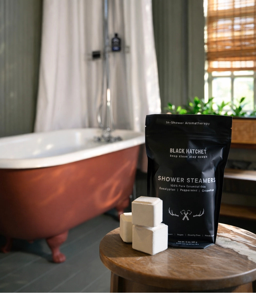 A black bag of Black Katchey Shower Steamers sits on a wooden table next to three white cube-shaped steamer blocks in a rustic bathroom setting with a copper tub and shower visible in the background.
