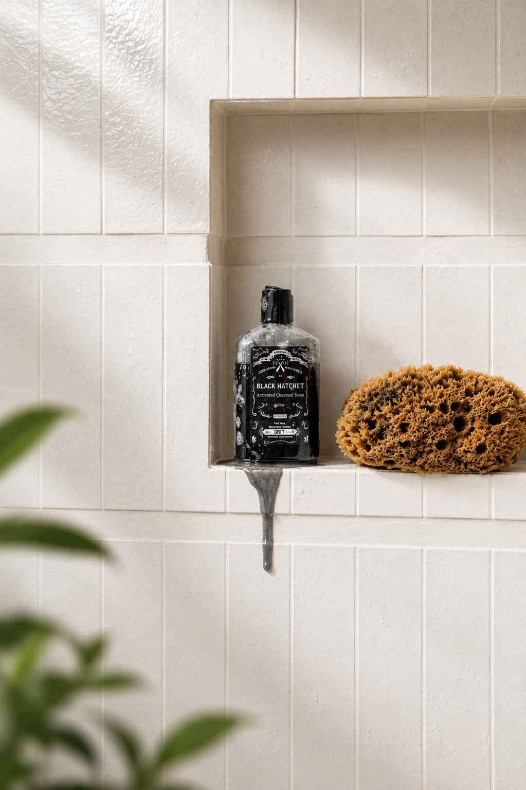 A bottle of Black Knight shower gel stands on a small metal drip tray in a recessed shelf of a cream tiled wall, next to a brown sponge. Blurred green leaves are visible in the foreground.