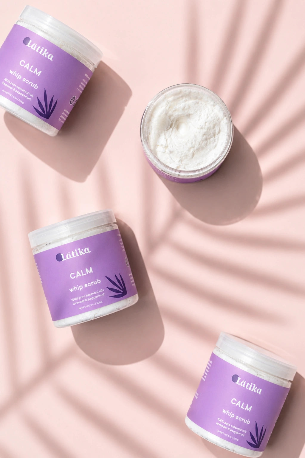 Four jars of Latika Calm Soap Scrub arranged on a soft pink background with leaf shadow overlays. Three jars are closed with lavender labels, and one open jar at the top right shows the white scrub inside.