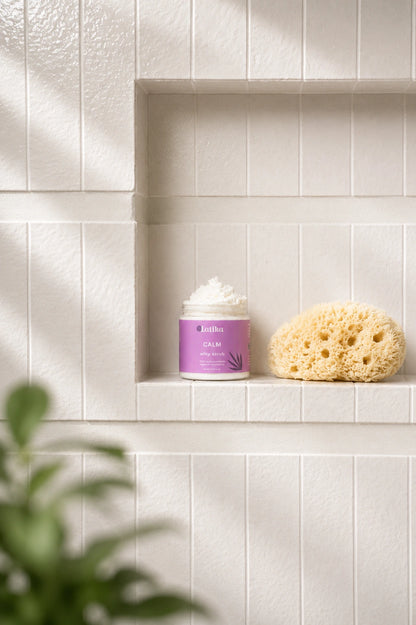 A jar of Bath &amp; Body Works Calm body cream with a purple label sits on a white tiled shelf next to a natural sponge, with soft sunlight and a blurred green plant in the foreground.