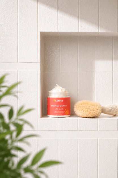 A jar of Luniko Energy Boost Body Scrub with whipped cream visible at the top, set on a white tiled shelf next to a natural bristle brush. A green plant leaf is visible in the foreground, and the background is a clean white tiled wall.
