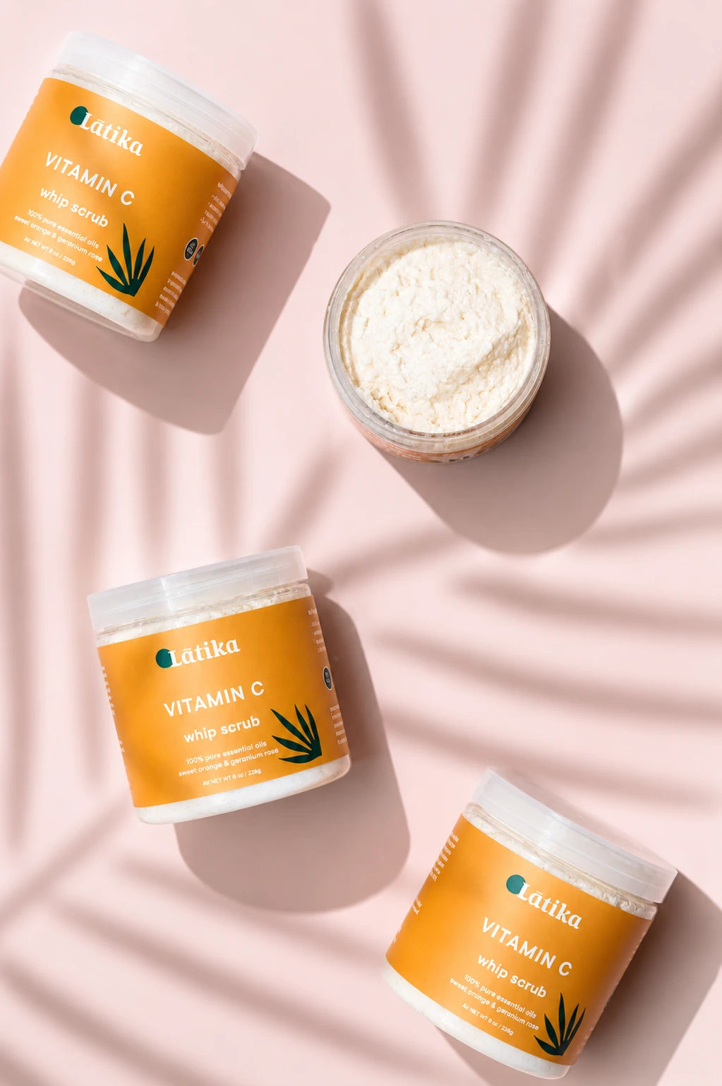 Three jars of Latika Vitamin C Whip Scrub arranged on a pink background with soft leaf shadows. Two jars are closed with orange labels, and one open jar shows the white scrub inside.
