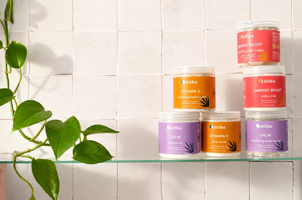 Five jars of Latika skincare products arranged on a glass shelf against a white tiled wall, with a green plant leaf trailing from the left side. The products include Energy Boost, Vitamin C, Calm, and other variants in orange, red, and purple packaging.