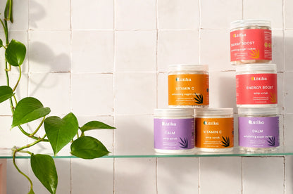 Five jars of Latika skincare products arranged on a glass shelf against a white tiled wall, with a green plant leaf trailing from the left side. The products include Energy Boost, Vitamin C, Calm, and other variants in orange, red, and purple packaging.