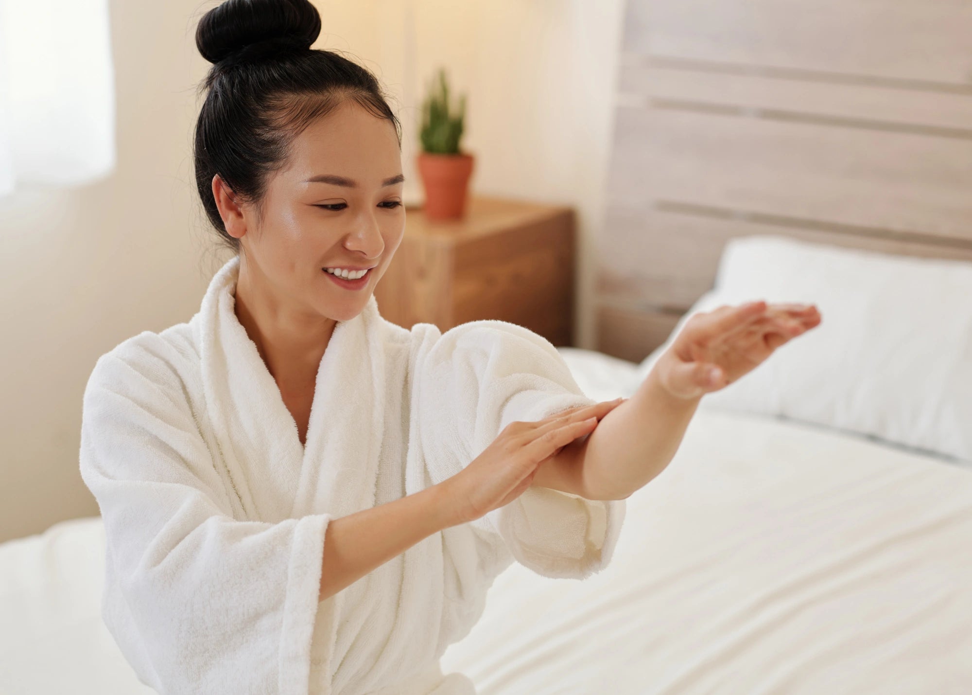 Woman in a white robe applying lotion to her arm in a bedroom setting.