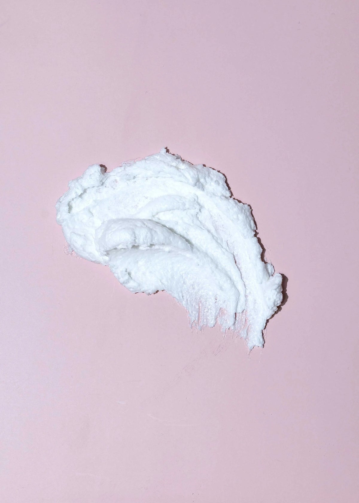 A dollop of thick white cream or balm with a swirled texture, placed on a solid light pink background.